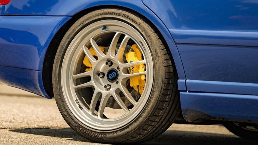 Close-up of a blue car's rear wheel featuring a silver multi-spoke alloy wheel, a low-profile tire, and bright yellow brake calipers.